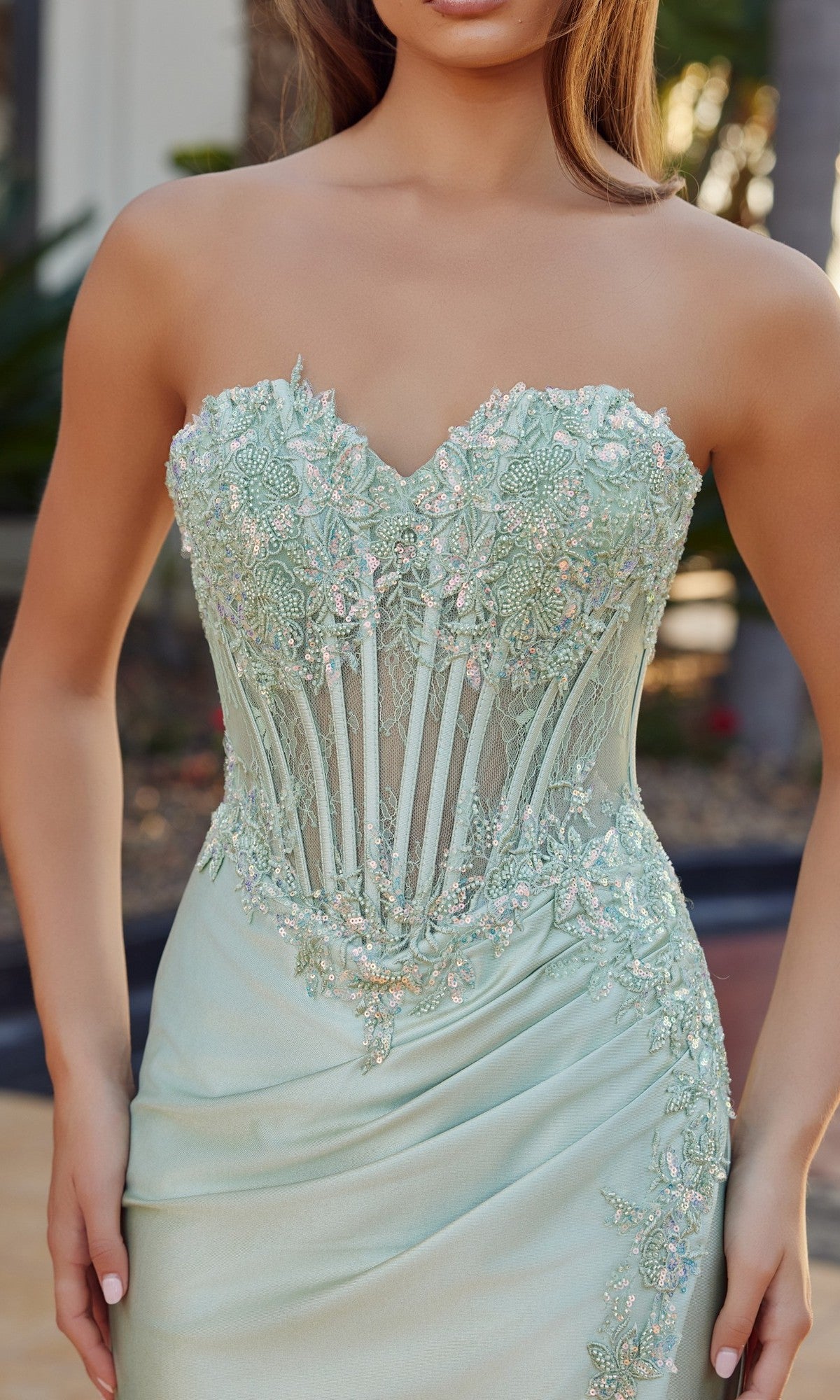 A woman stands outdoors in the Nox Anabel R1811 long prom dress—a strapless satin gown in mint green with a high-leg slit, floral lace embroidery, and an embroidered corset bodice.
